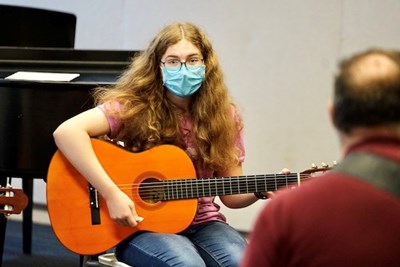 A girl in glasses and a face covering holds an acoustic guitar while looking at a teacher