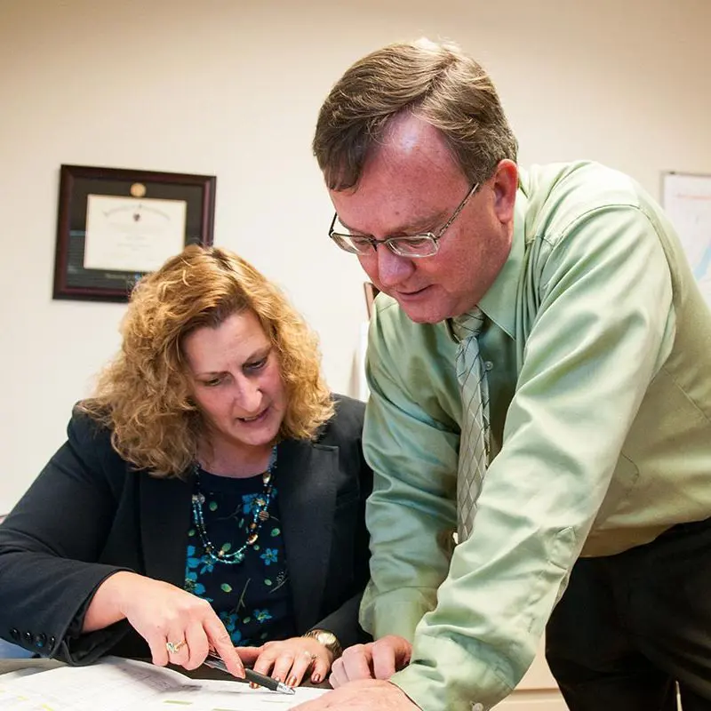 Two employees at desk looking down at paperwork and one pointing at something.