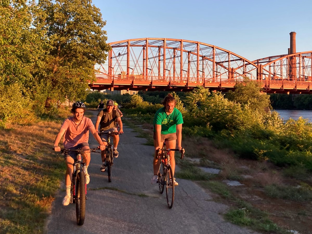 three people ride a bike in the warm sunset glow with the Aiken St bridge behind