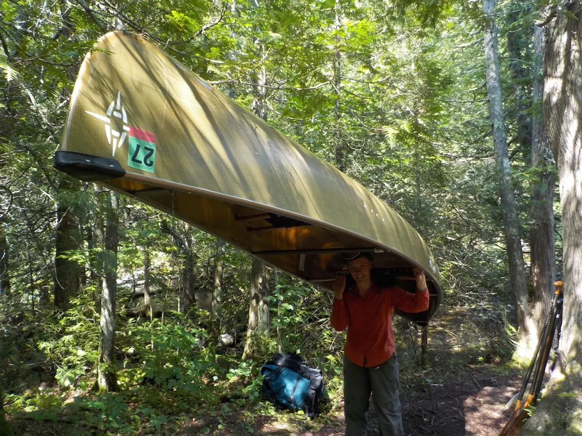 A person carries a canoe upside on her shoulder and smiles.