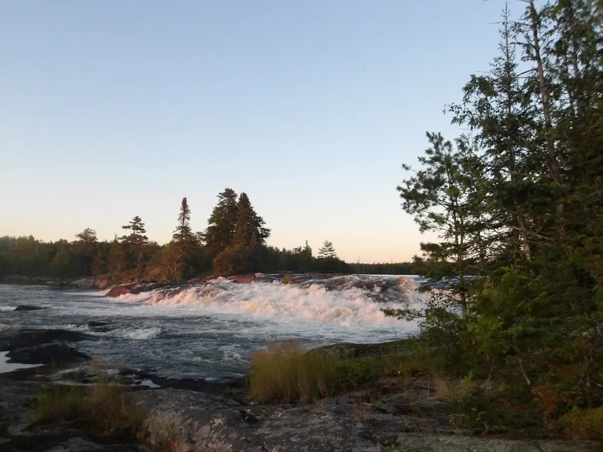 A waterfall flows across and past a rocky shoreline with a few trees on either side.