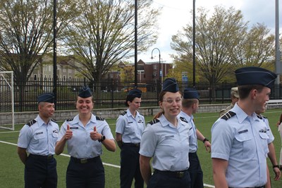 A group of AFROTC cadets standing in a line outside on the UMass Lowell campus. One giving a double thumbs up symbol.