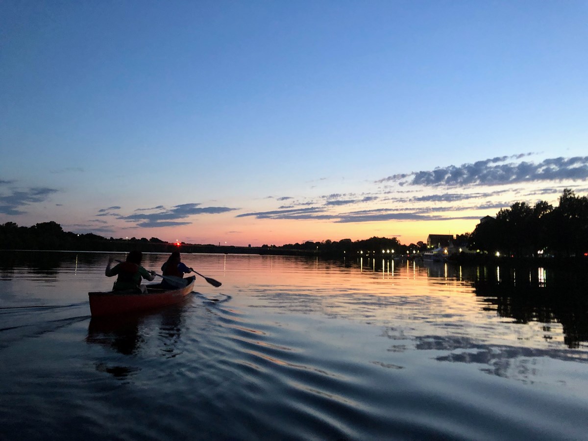 Two people paddle a canoe at sunset with a darkening sky.