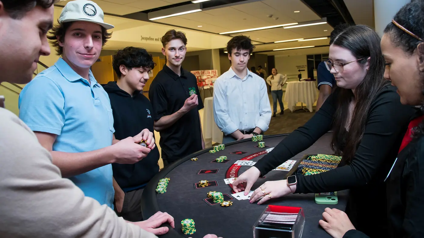 Young men stand around a casino table where young women deal cards