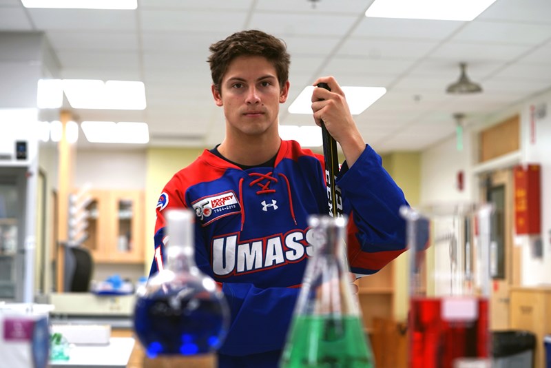 Charlie Levesque stands with his hockey stick in uniform in a chemistry lab, blurred out beakers of colored liquid in the foreground