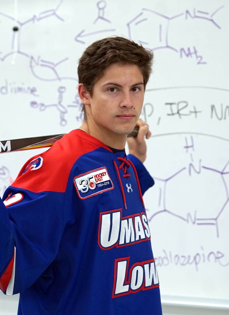Charlie Levesque stands in front of a whiteboard with molecules drawn on it in his hockey uniform with his hockey stick over his shoulders