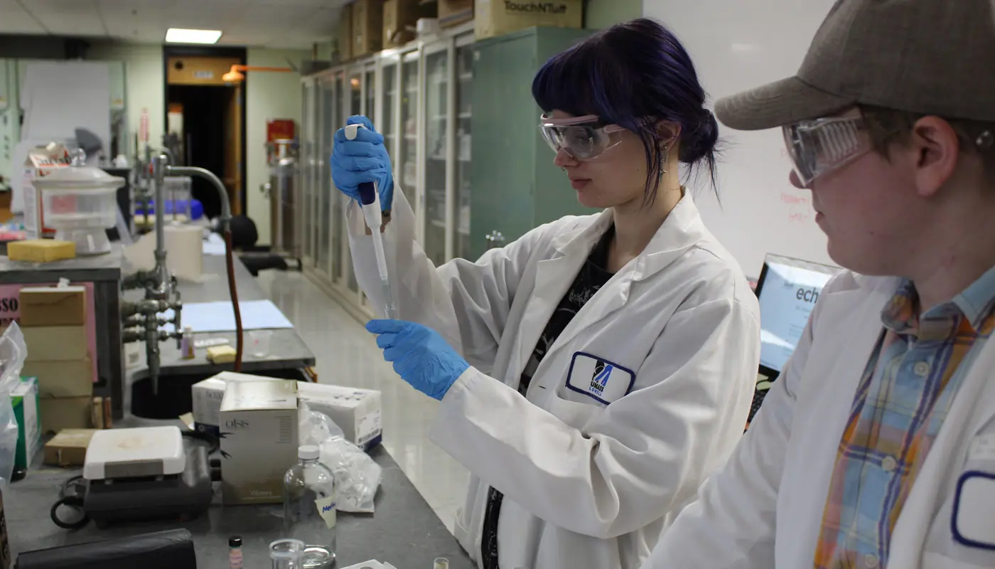 A student adds liquid to a test tube while another student watches.