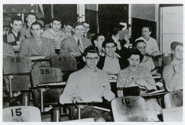 Students sit in a lecture hall in the 1950s in wooden numbered chairs
