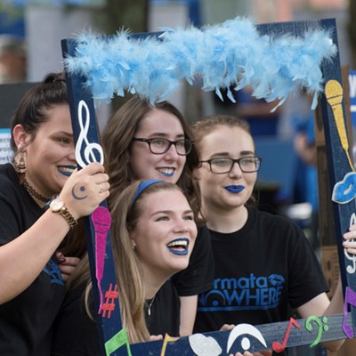 Four female students wearing blue lipstick pose in a handmade frame decorated with musical notes at the UMass Lowell Club Fair