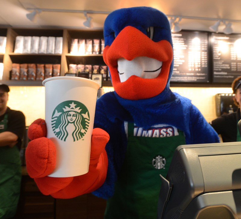 Rowdy the River Hawk hoding a coffee cup and wearing a green apron behind the cash register at Starbucks on North campus