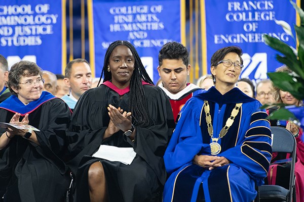 A woman in a black robe applauds while seated on a stage with people in academic regalia
