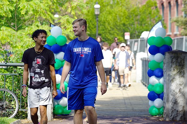 Two smiling students walk past balloons while students cross a bridge behind them