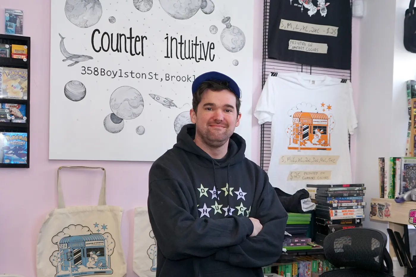 A young man in a black hoodie and baseball cap poses for a photo with his arms folded inside a record store.