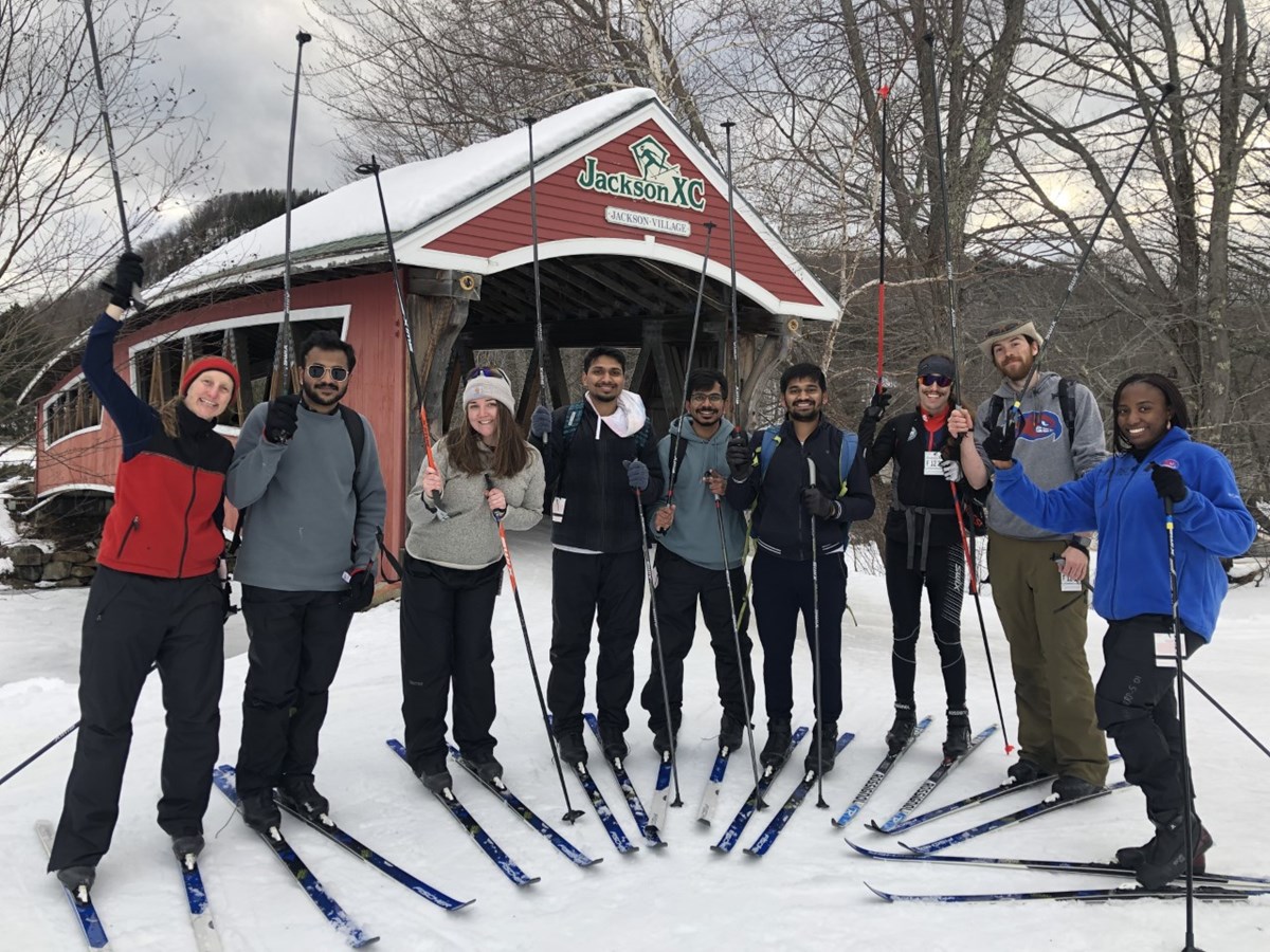 a group of nine people on cross country skis poses in front of a covered bridge, all smiling and holding up their ski poles