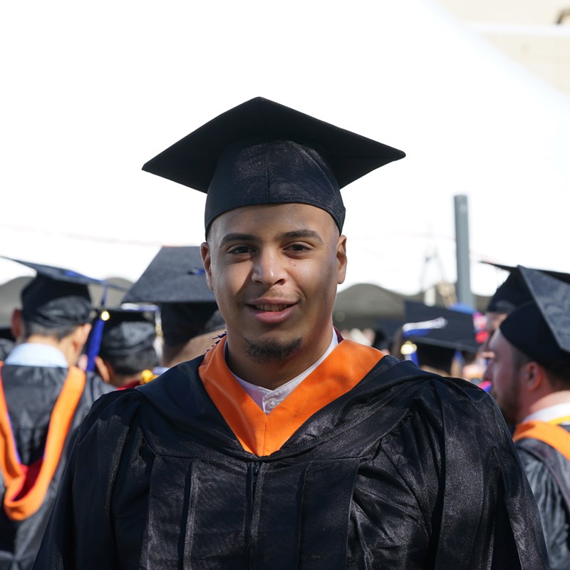 Headshot of Dayer Acosta in a cap in gown outside the Tsongas Center at UMass Lowell at Commencement 2019