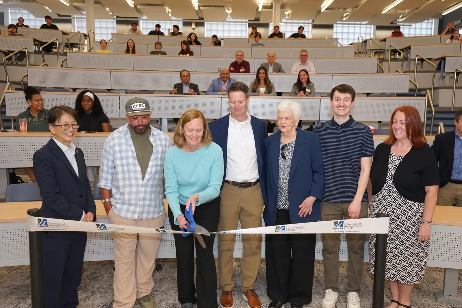 A woman cuts a white ribbon with giant scissors while six people look on in a college lecture hall.