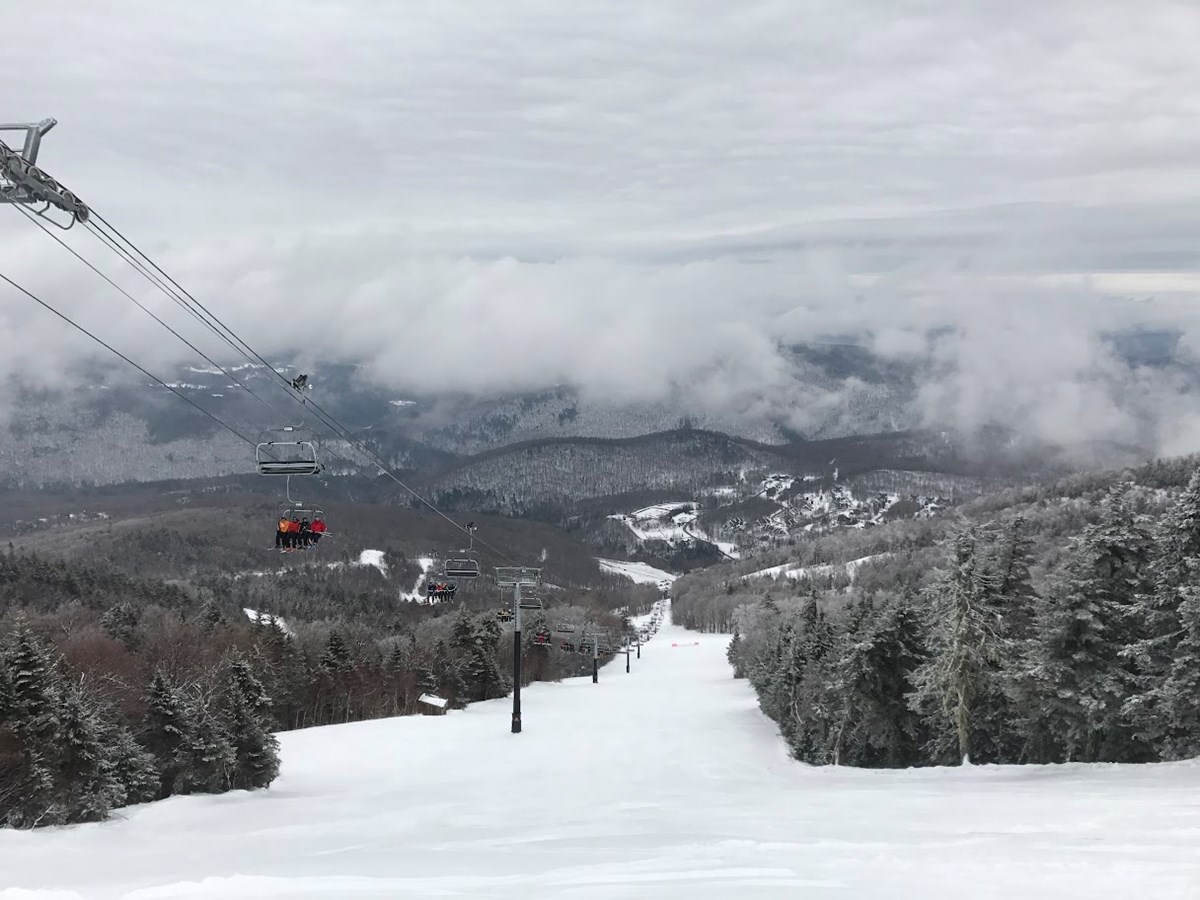 a wide ski trail leads to a view of a mountains with ski trails visible beyond as low clouds hang on the side