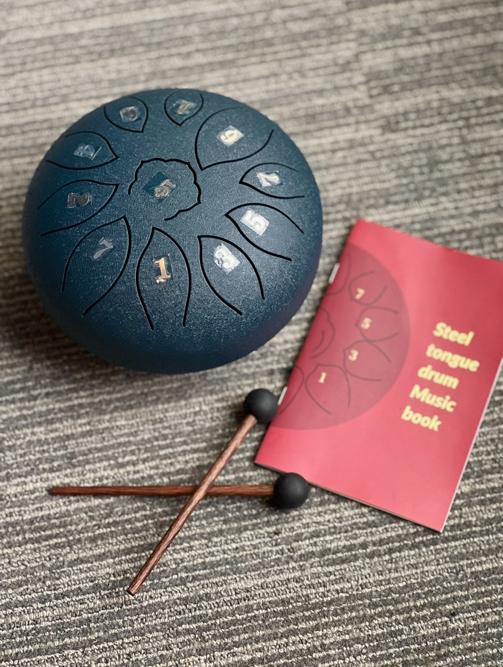 Blue drum labeled with numbers 1-6, two wooden drum mallets with a black rubber top and a maroon steel tongue drum music book.