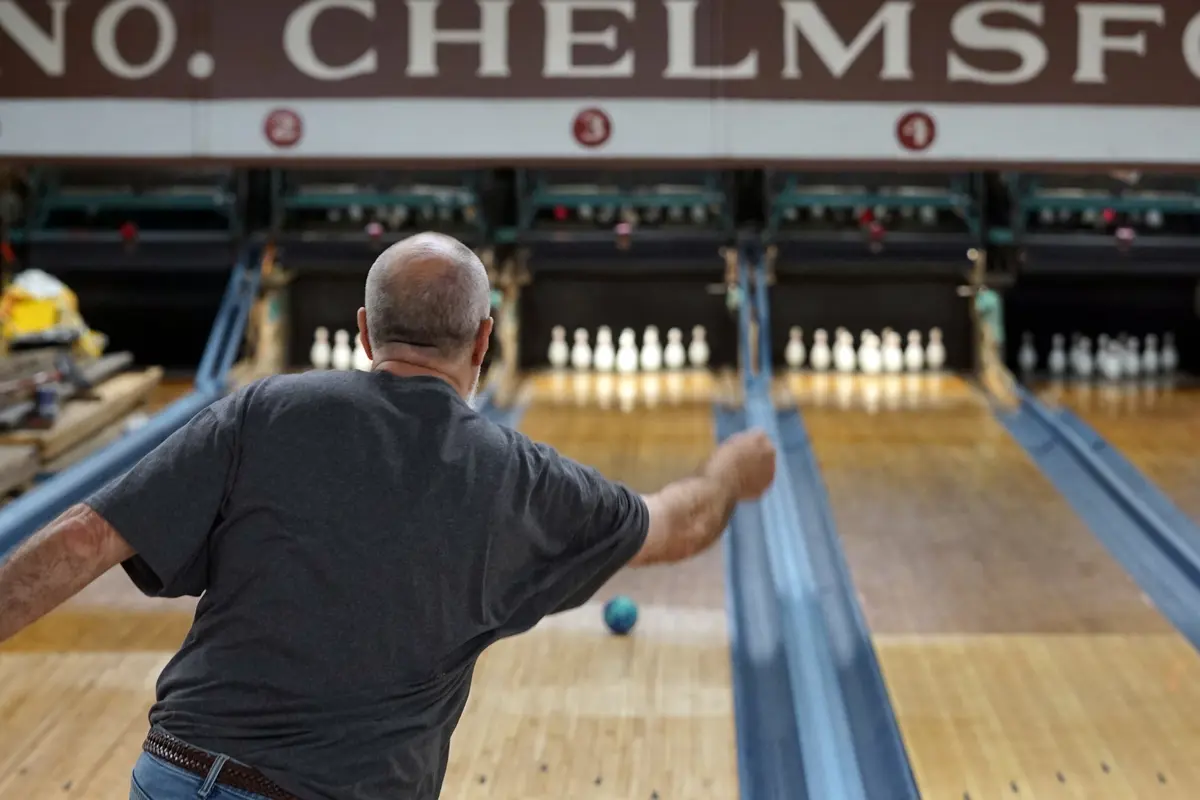 A man watches a duckpin bowling ball travel down the lane toward the pins.