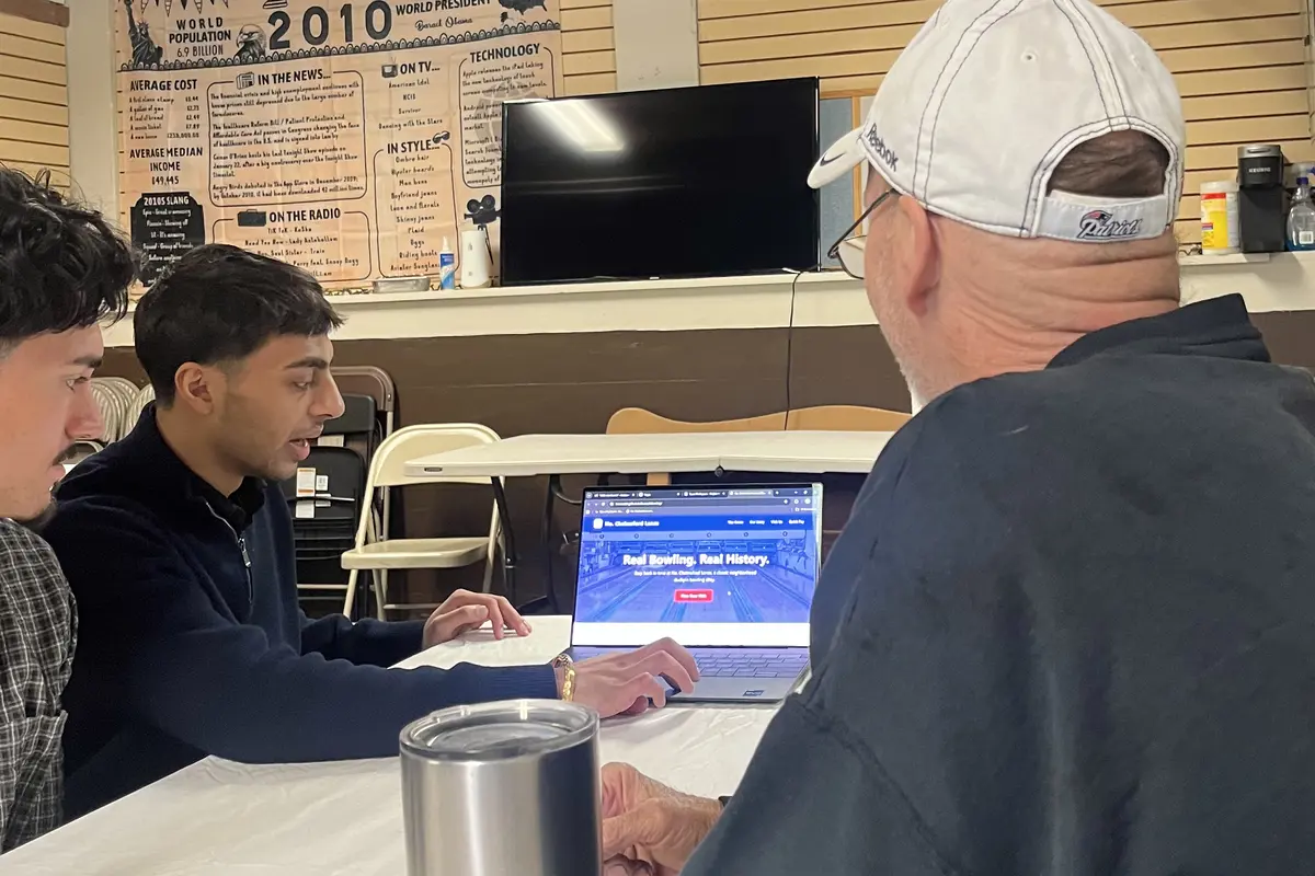 A young man with dark hair shows a man in a white hat a laptop screen while another young man looks on.