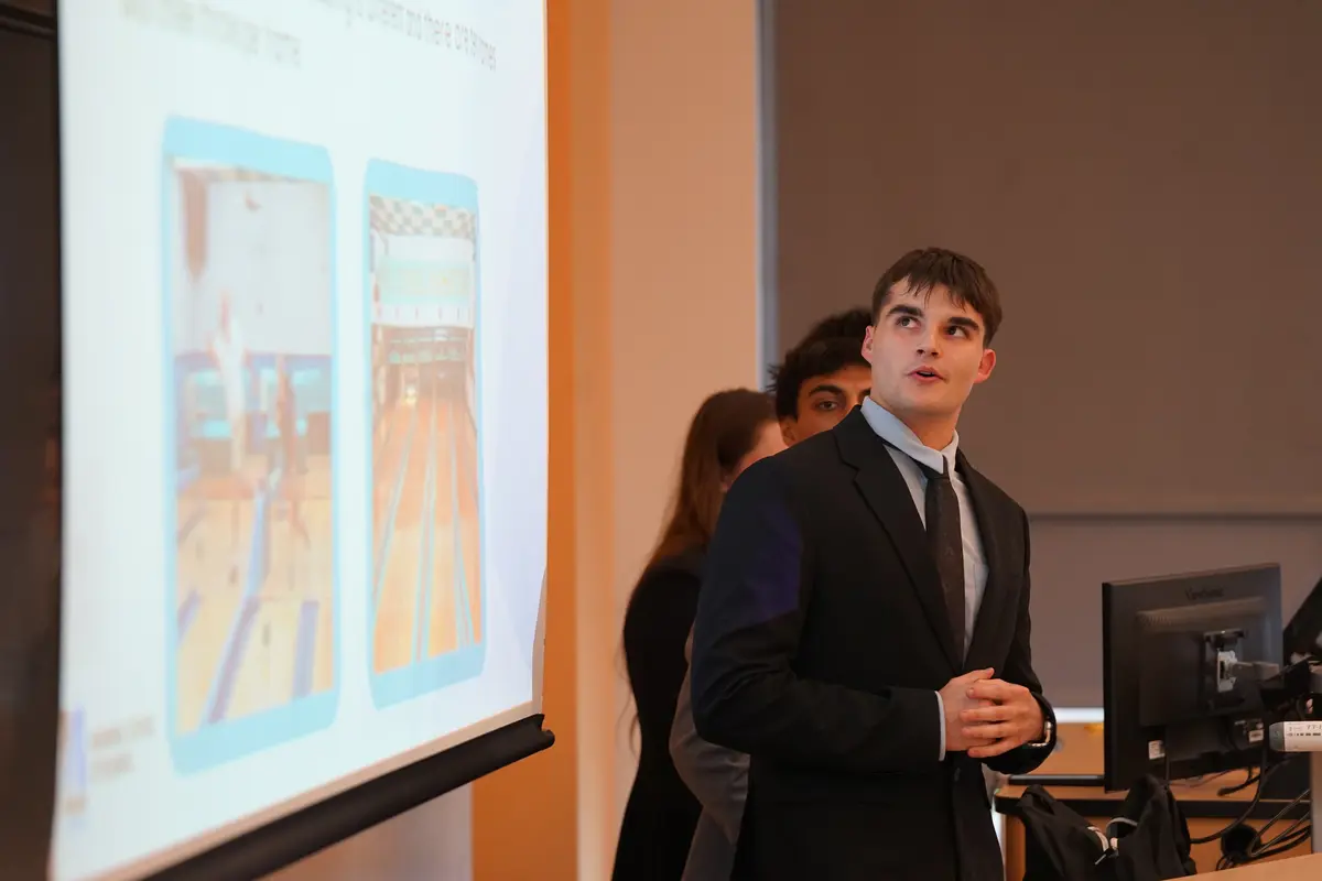 A young man in a suit looks back at a projection screen while giving a presentation.