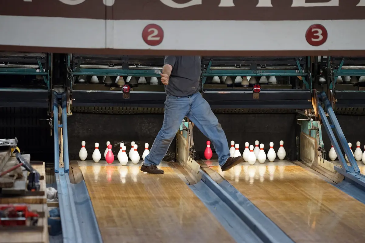 A man give a thumbs up sign while walking across duckpin bowling lanes.