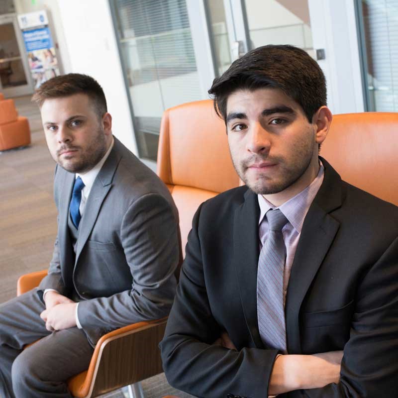 UMass Lowell students and Model U.N. members Alejandro Lopez and Ryan Dekeon, pictured in suits sitting in the Pulichino Tong Building in orange chairs looking at the camera