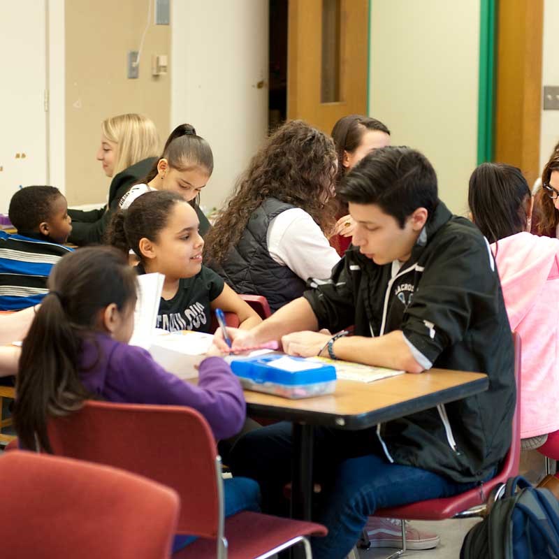 A UMass Lowell education student sits at a table with two young students in a school classroom