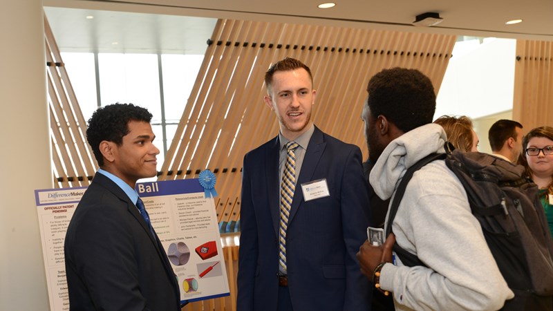 Edward Morante (left) stands with Benjamin McEvoy in front of their poster at a DifferenceMaker event, talking to a student