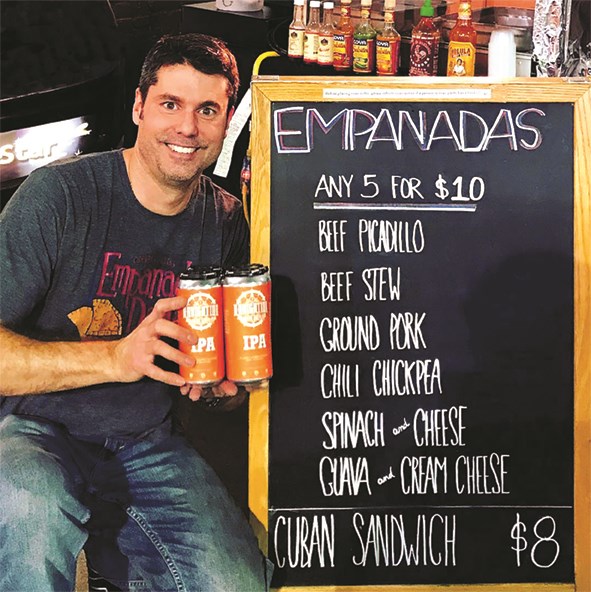 A man holds up cans of IPA in front of the chalkboard menu at Empanada Dada