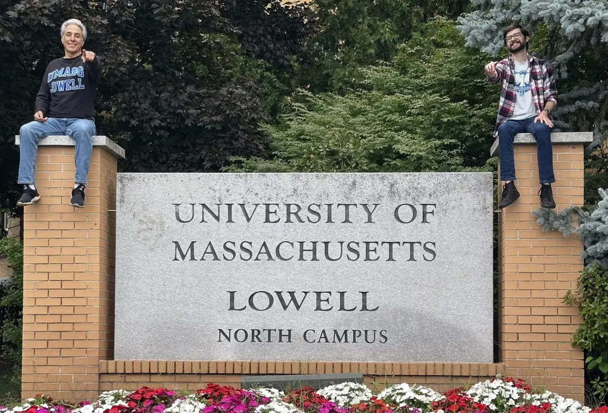 Two men pose for a photo while sitting on top of pillars for a college sign outside.