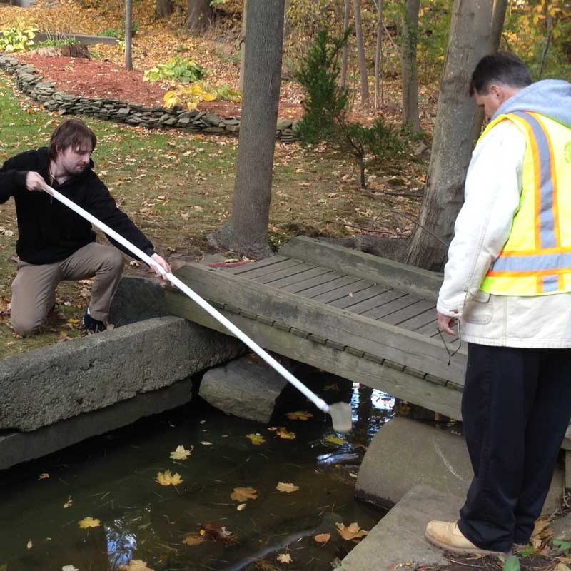 A UMass Lowell environmental student holds a white pole with a cup over a small stream to take a water sample.