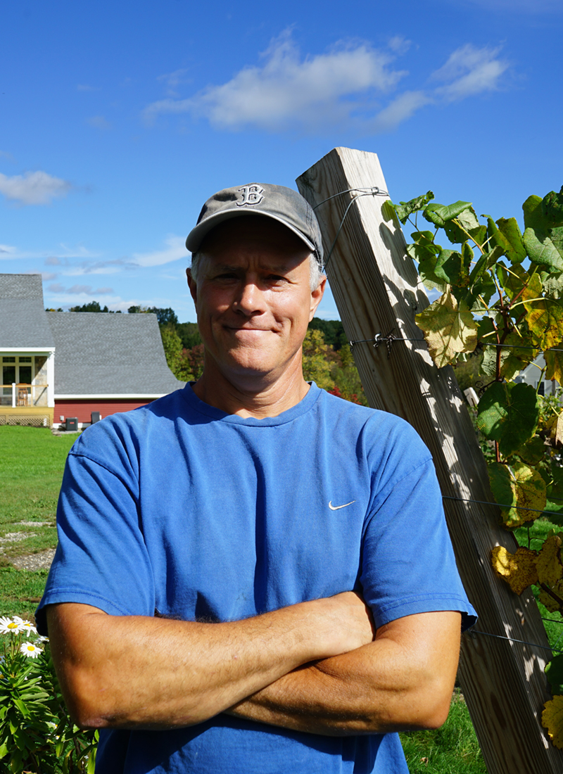 Eric Preusse stands in between rows of grapes on a sunny day at his vineyard, Broken Creek