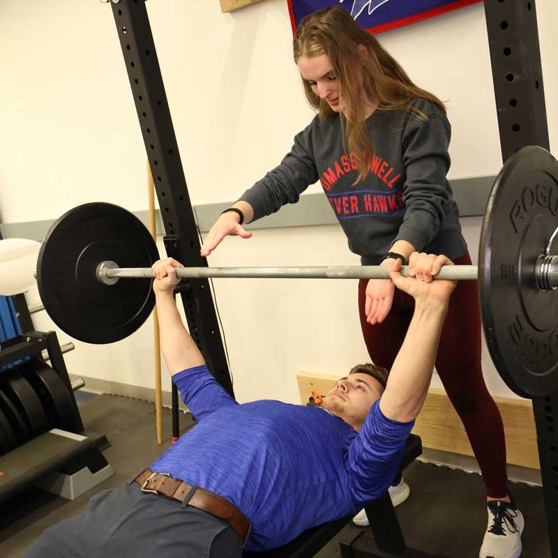 One student student lifts a weight as another student spots in a UMass Lowell exercise science facility