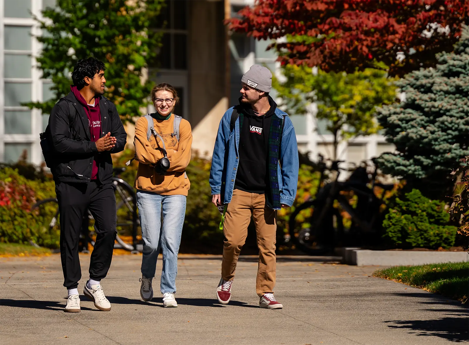 Three people walking in front of shrubbery and trees with a building in the background