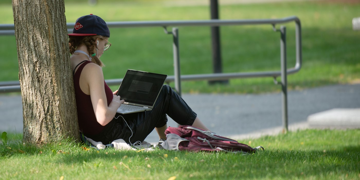 Female student sitting up against a tree working on her computer