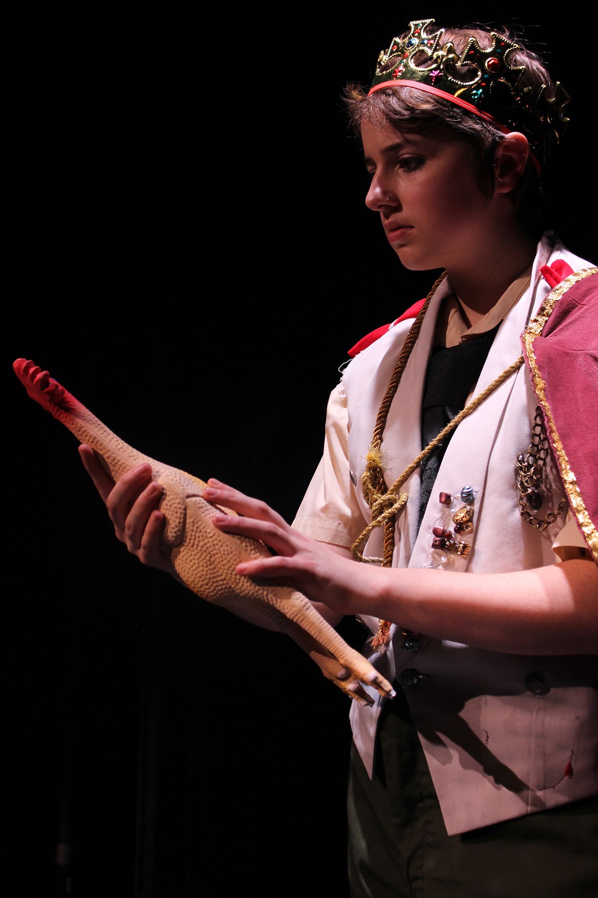 A female student wearing a crown and holding a rubber chicken on stage during a UMass Lowell Theatre Arts production.
