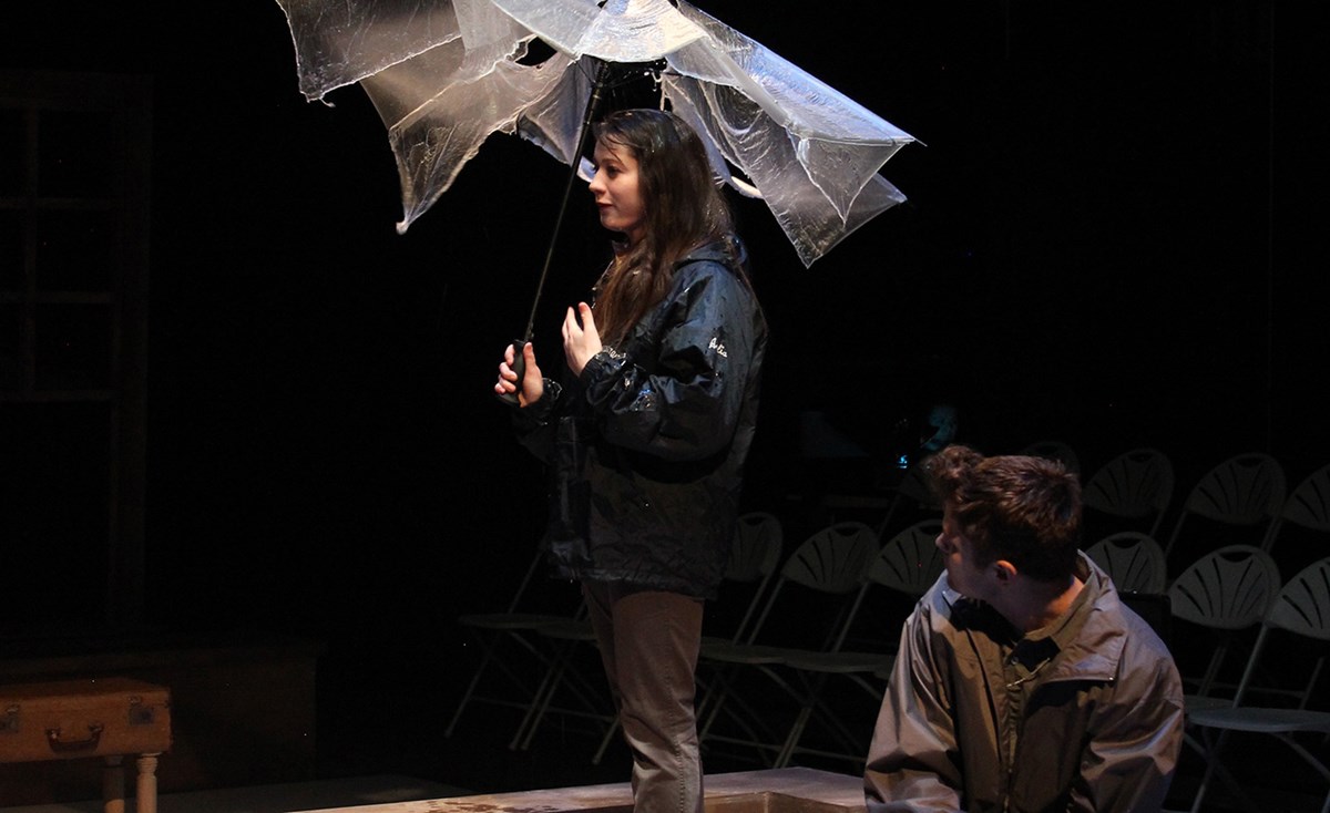 A female holding an umbrella on stage while a male student looks on during a UMass Lowell Theatre Arts production.