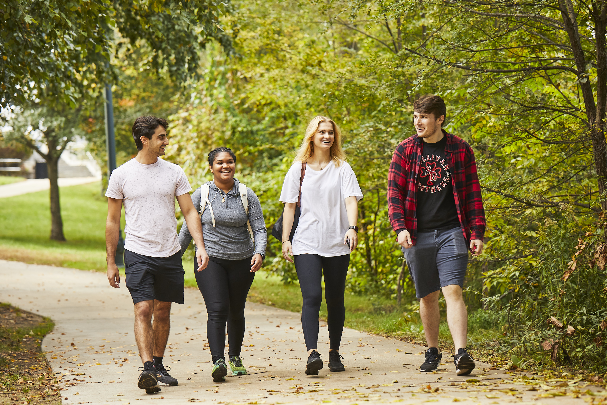 Four UMass Lowell students (2 male and 2 female) smiling and walking down a wooded path on campus.
