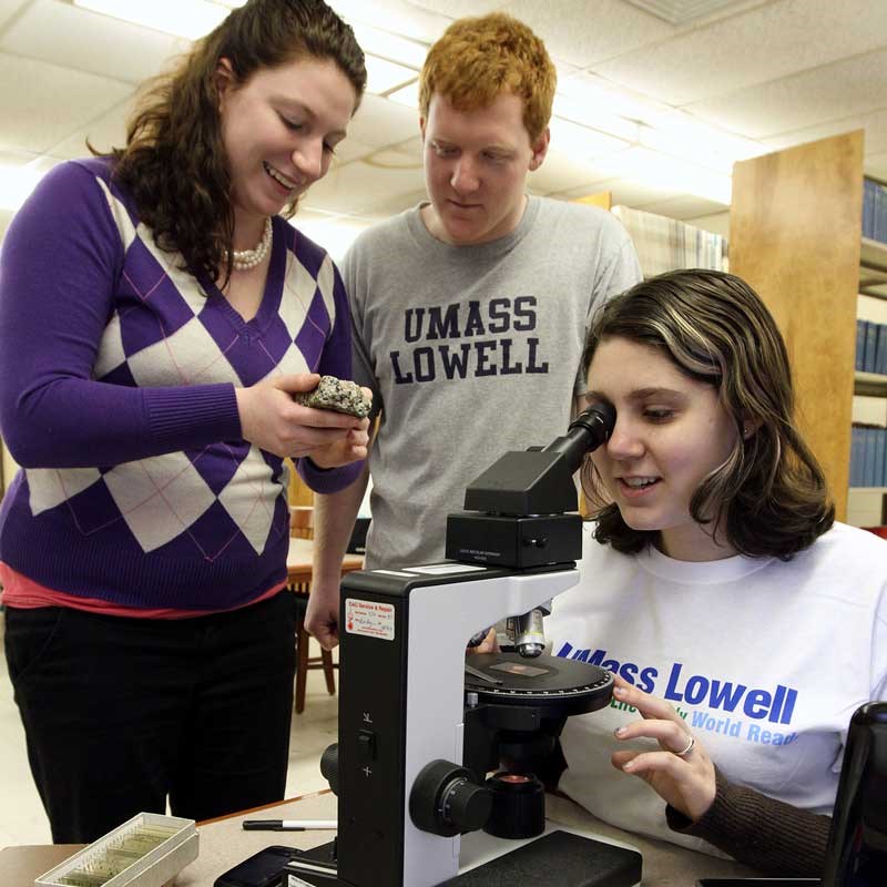 UMass Lowell environmental science student looks through a microscope as two other students stand nearby looking at a rock sample