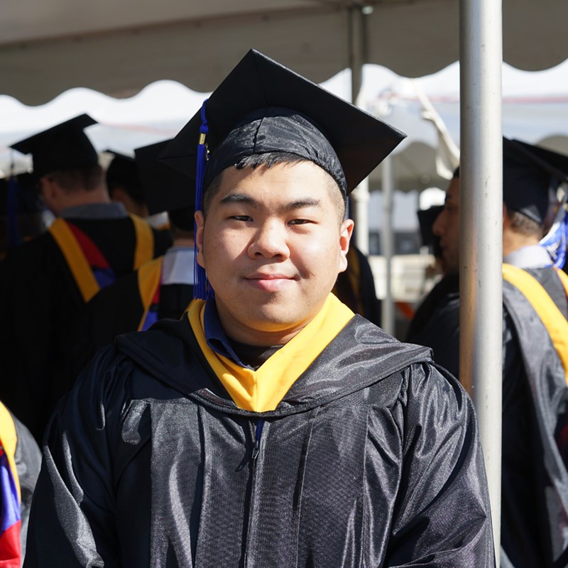 Headshot of George Le in a cap in gown outside of the Tsongas Center at UMass Lowell at Commencement 2019