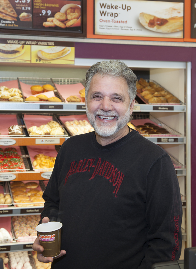 George Zografos holds a cup of coffee in front of racks of donuts at one of his former Dunkin' Donuts locations on Cape Cod