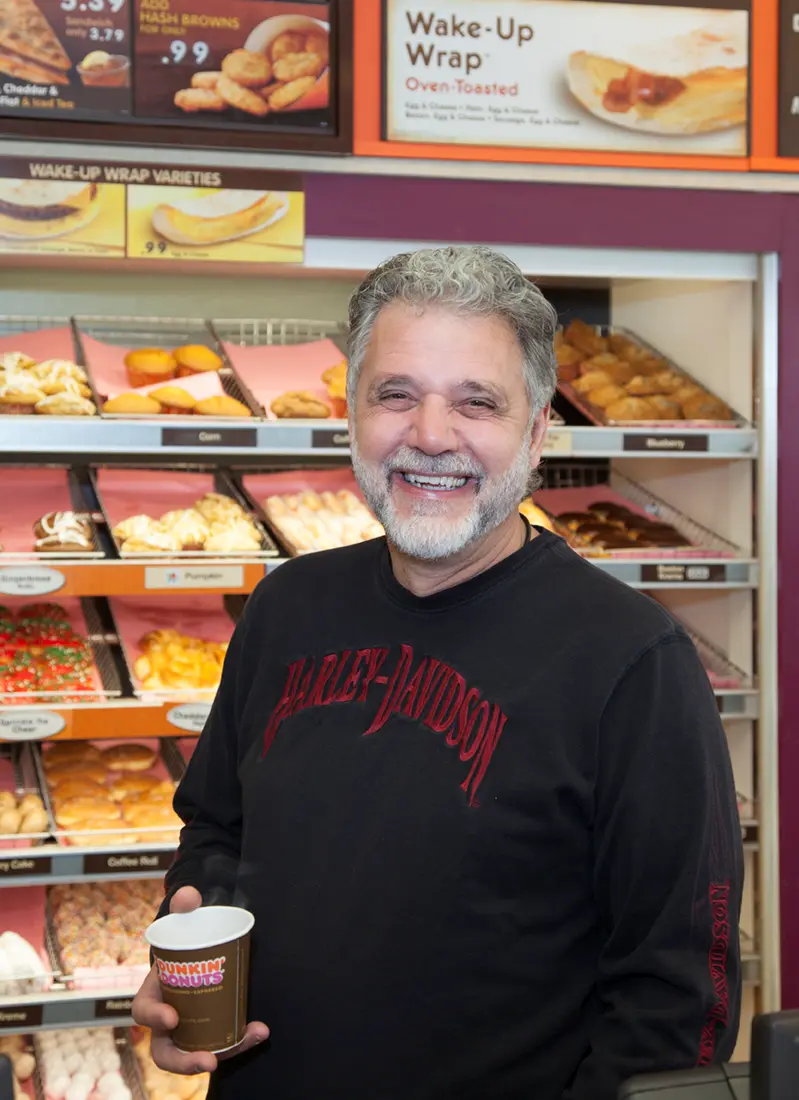 George Zografos holds a cup of coffee in front of racks of donuts at one of his former Dunkin' Donuts locations on Cape Cod