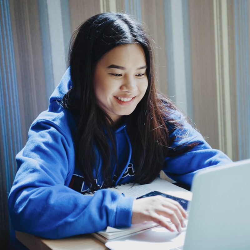 female student in blue hoodie sitting at a table with a laptop
