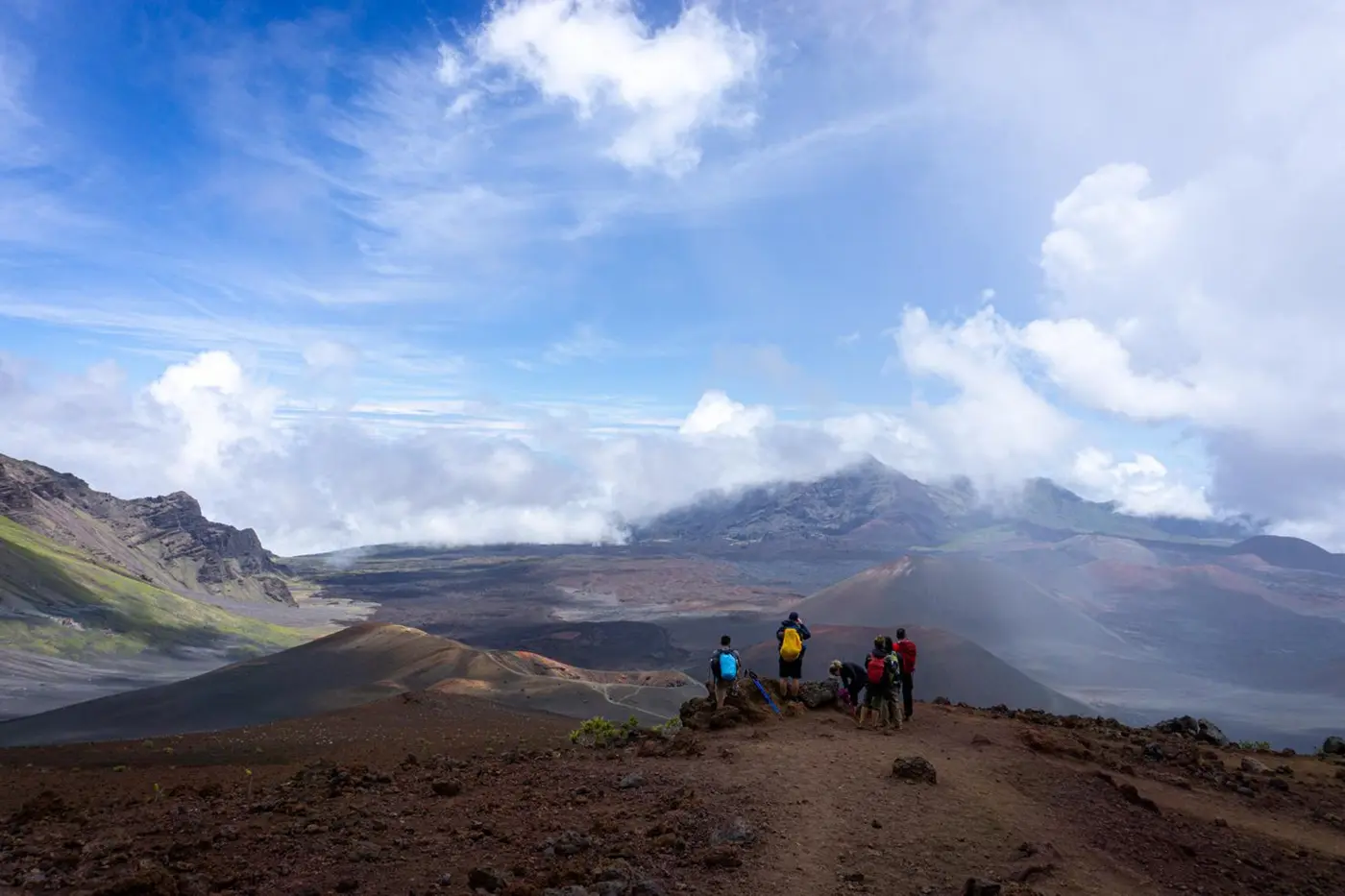 A group of 5 pepole with colorful backpacks stand looking far away at a rocky scene with mist rising. 