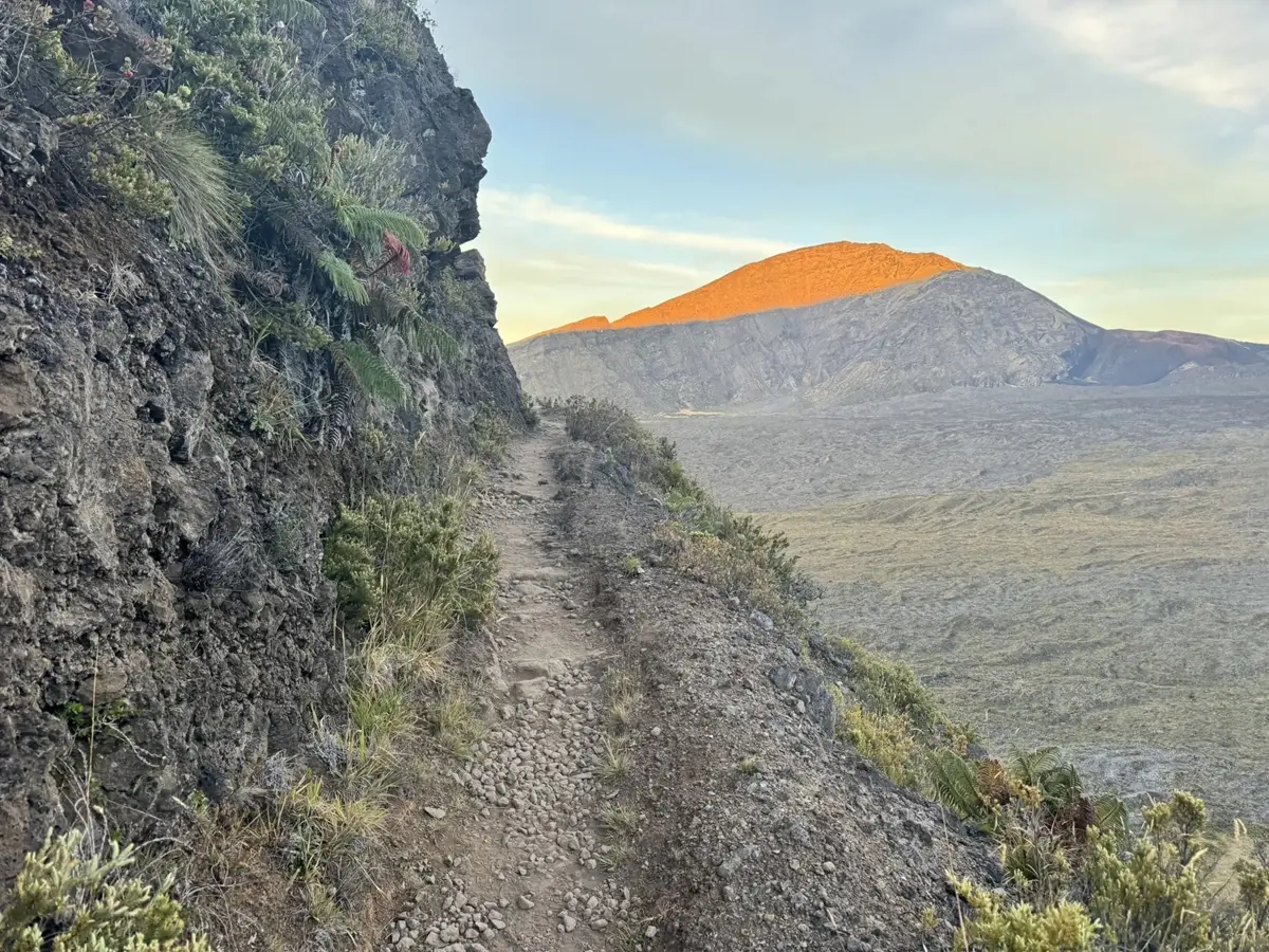 A trail leads past a steep drop off with a sunlight mountain beyond.