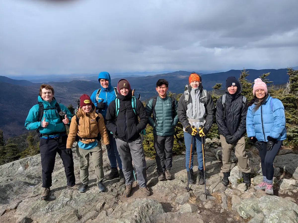 A group of eight people in warm layers stands atop a bar summit with clouds beyond.