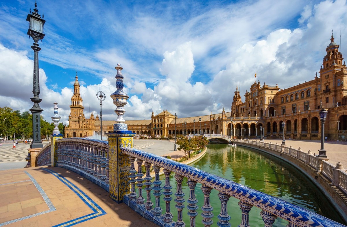 Bridge going over a river with the Plaza de España in Seville, Spain and a cloudy blue sky in the background.