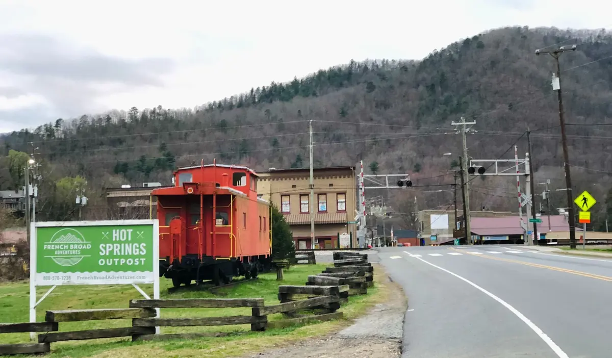 A sign says "Hot Springs Outpost" as the road enters into the town with a small rail road car, buildings, and a tall hill. 