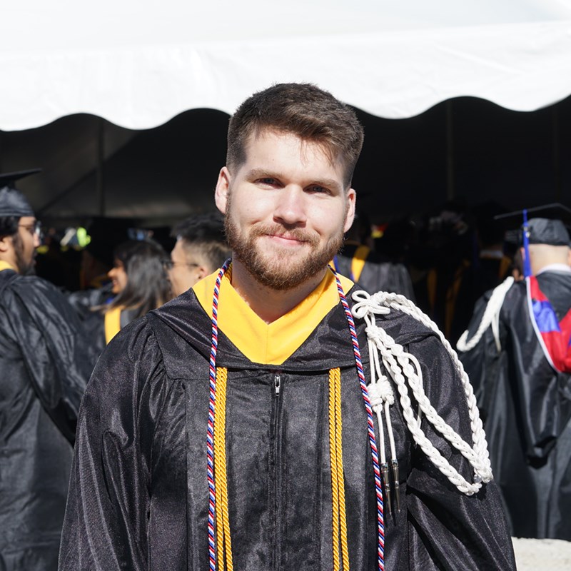 Headshot of Ian Connell in a graduation gown outside of the Tsongas Center at UMass Lowell at Commencement 2019
